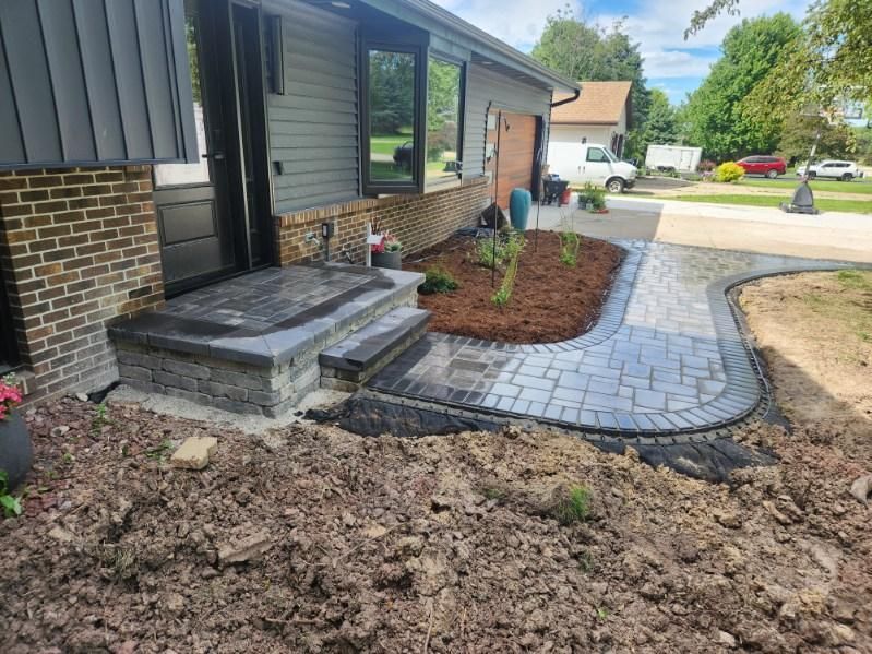 Exterior house with brick steps, paved path, and landscaping. Gray siding, black door, and cloudy sky.
