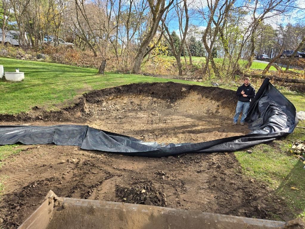 Man stands in excavated pond. Black liner partially covers the dirt. Green grass and trees surround.