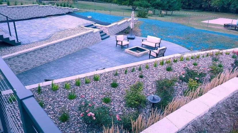 Patio with seating, fire pit, and landscaping, surrounded by retaining walls and a blue lawn.