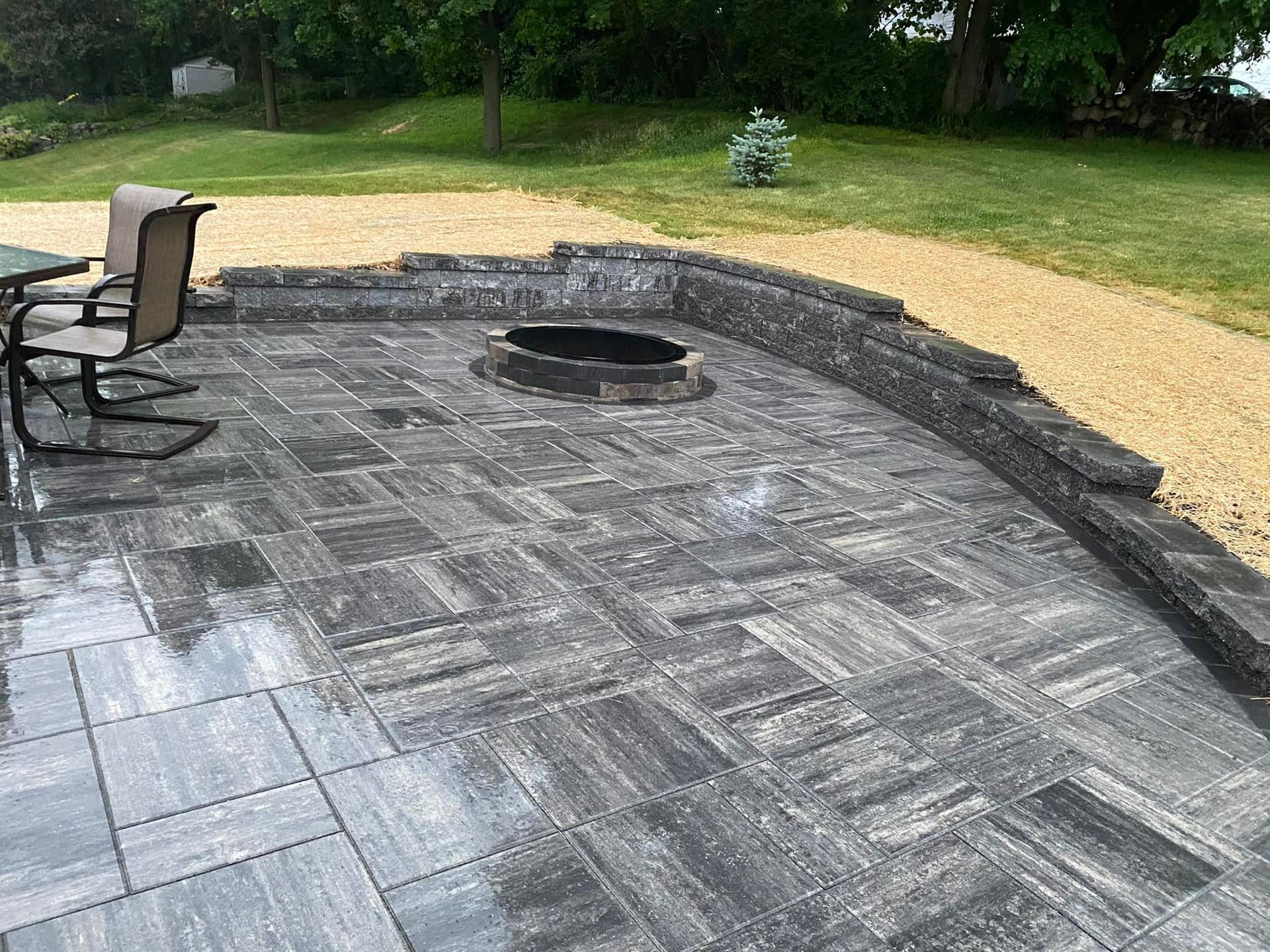 Patio with a fire pit, brick pavers, and gravel border. Two chairs sit at a table. Green grass in background.