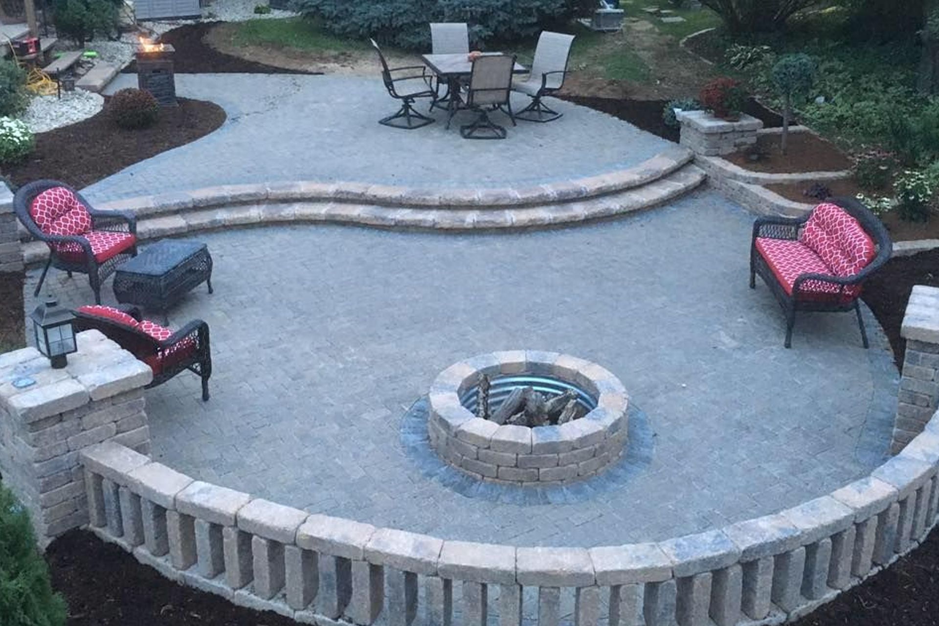 Outdoor patio with fire pit, seating, and dining area, surrounded by stone walls and landscaping.