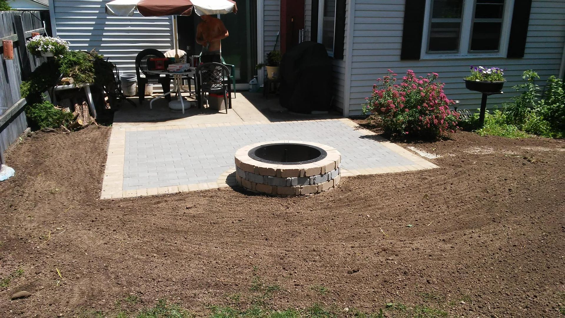 A backyard patio with a fire pit surrounded by mulch and a white house in the background.