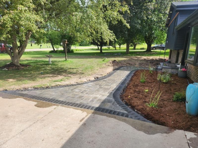 Brick pathway curves from a driveway, bordered by mulch and small plants, to a house entrance.