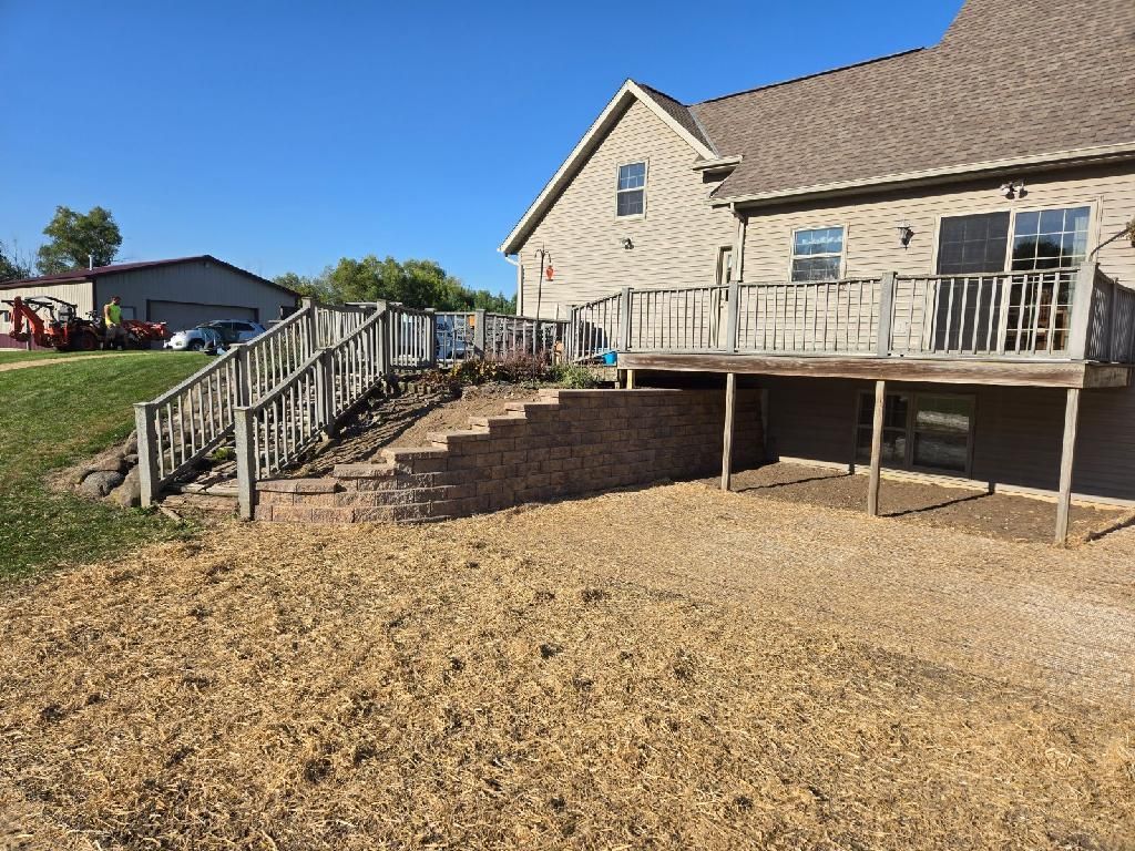 House with a deck, stairs, and retaining wall. Gravel area in front. Sunny day.