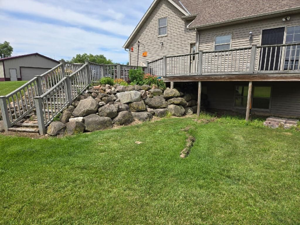 Backyard with a rock wall supporting a deck and stairs; green grass, blue sky.