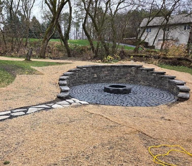 Stone fire pit with seating area in a gravel-covered yard near a house and trees.