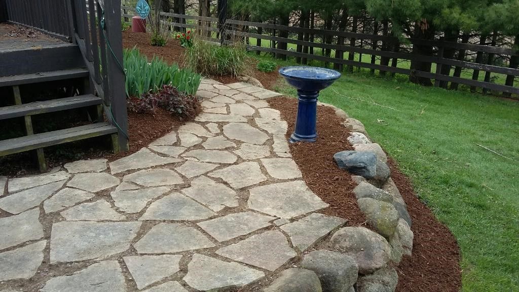 Stone path through garden bed, leading to birdbath. Steps, lawn, and wooden fence in the background.