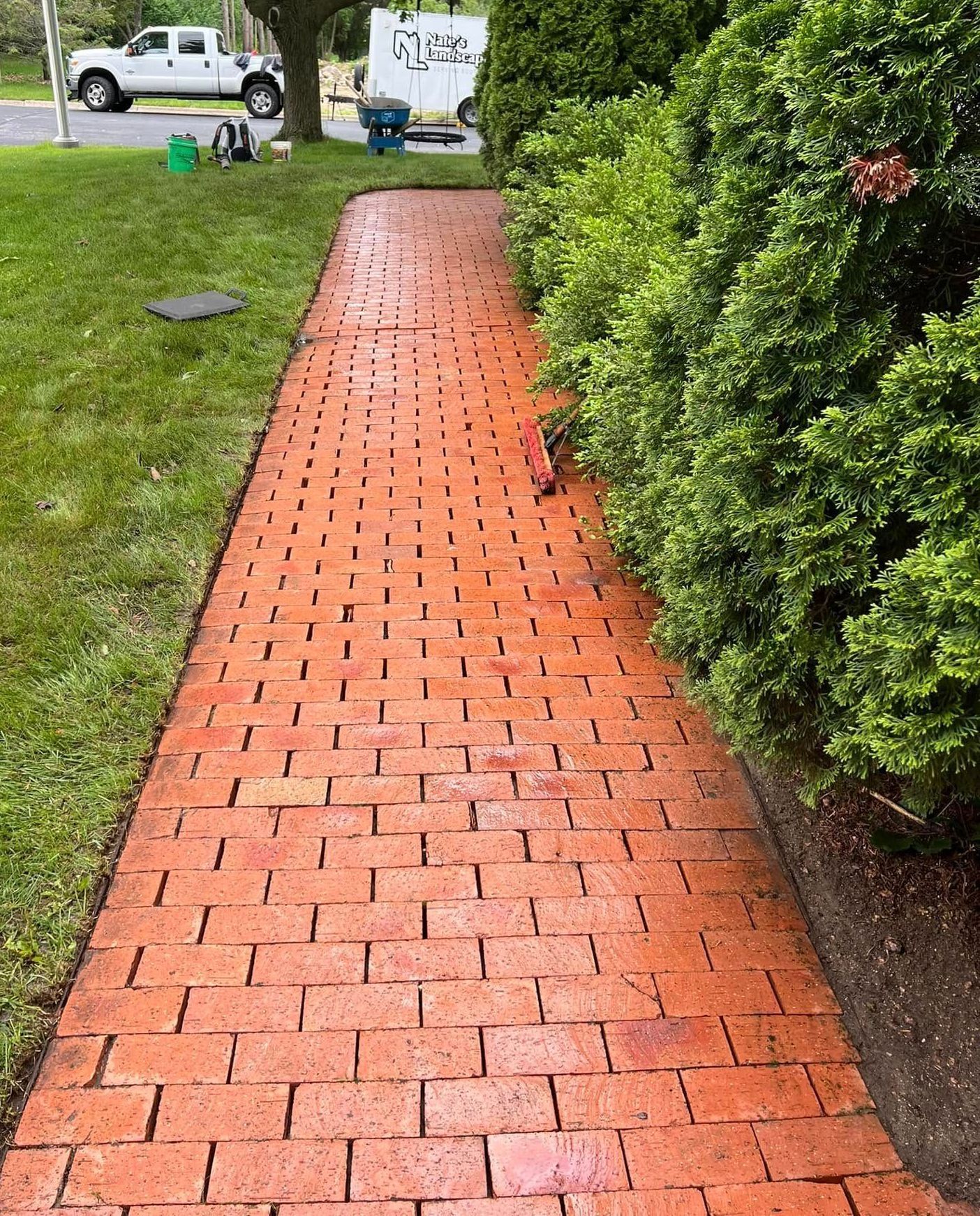 Brick walkway lined with green bushes and lawn. A white truck is in the background.
