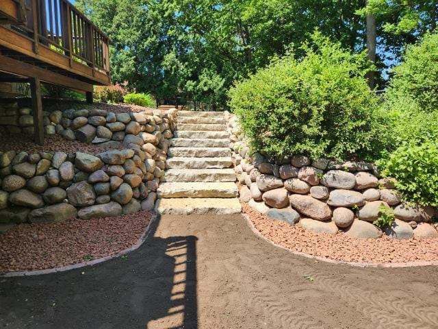 Stone steps and retaining walls lead up to a wooden deck, surrounded by greenery and red gravel.