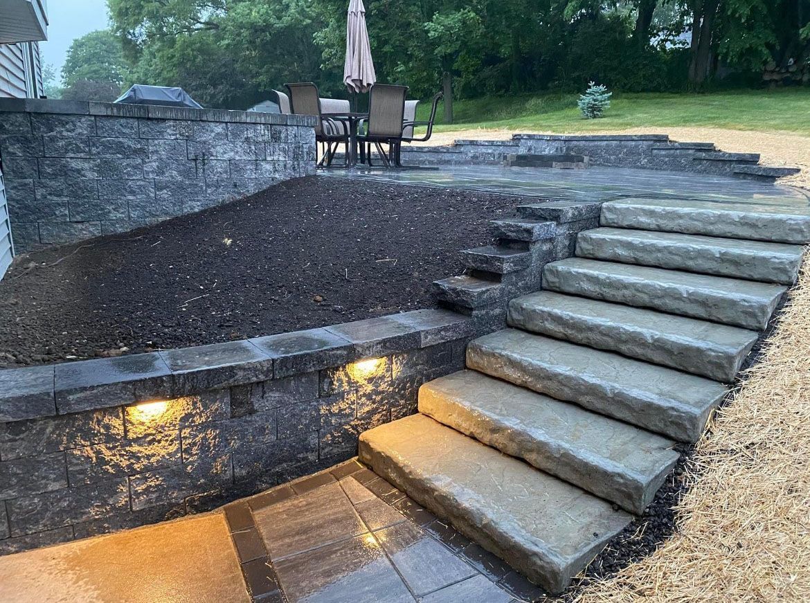Stone steps leading to a patio with a table and chairs. Dark stone retaining walls, lit with soft lights.