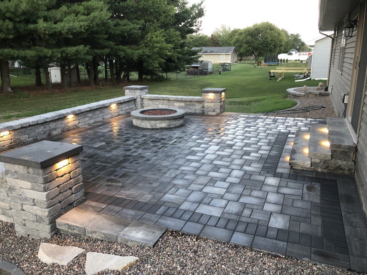 Brick patio with fire pit and built-in seating area, lit by outdoor lights at dusk.