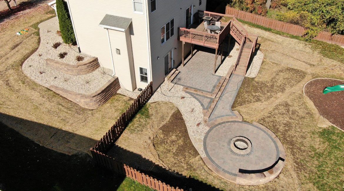 Aerial view of a house with a stone patio and fire pit in the backyard. Brown wood deck and retaining walls.