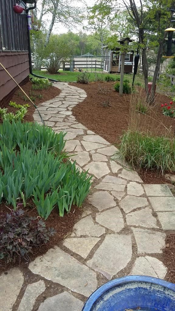 Flagstone path winds through a garden with green plants and mulch, leading toward a body of water.
