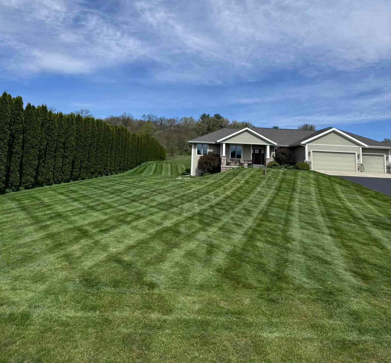 Green lawn mowed with stripes in front of a light green house, surrounded by trees, blue sky.