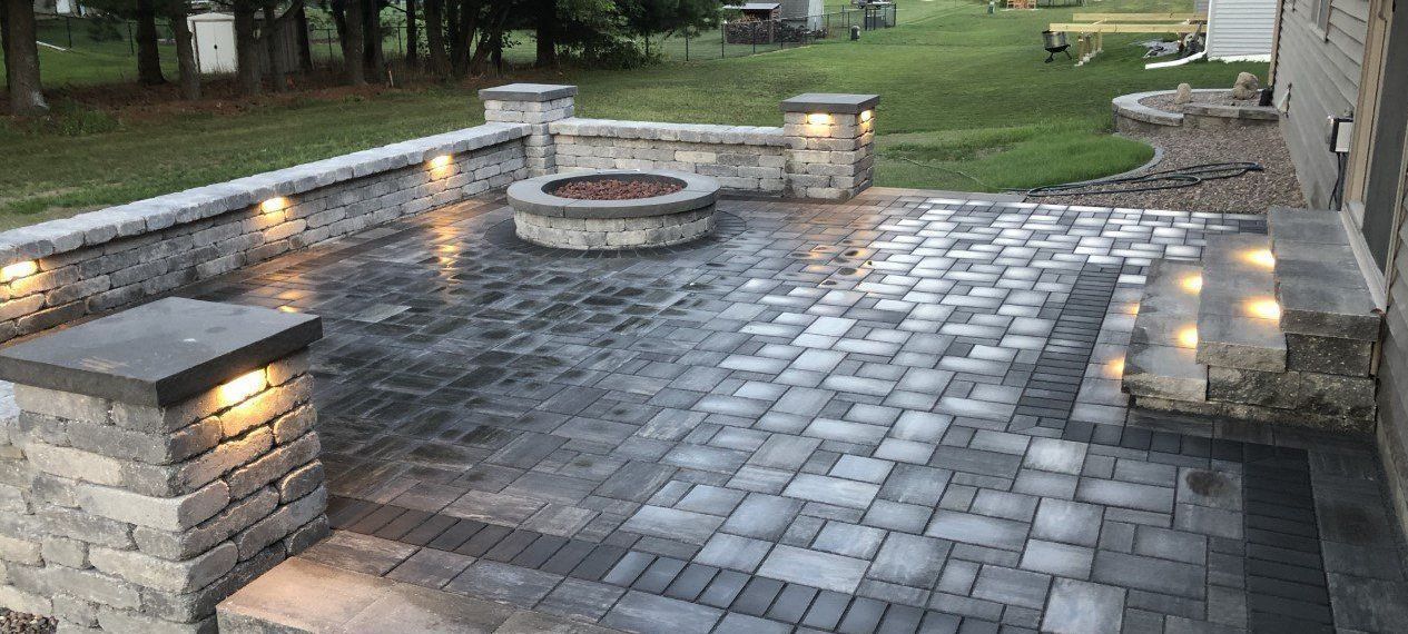 a brick patio with a fire pit, surrounded by a stone wall and posts with lights