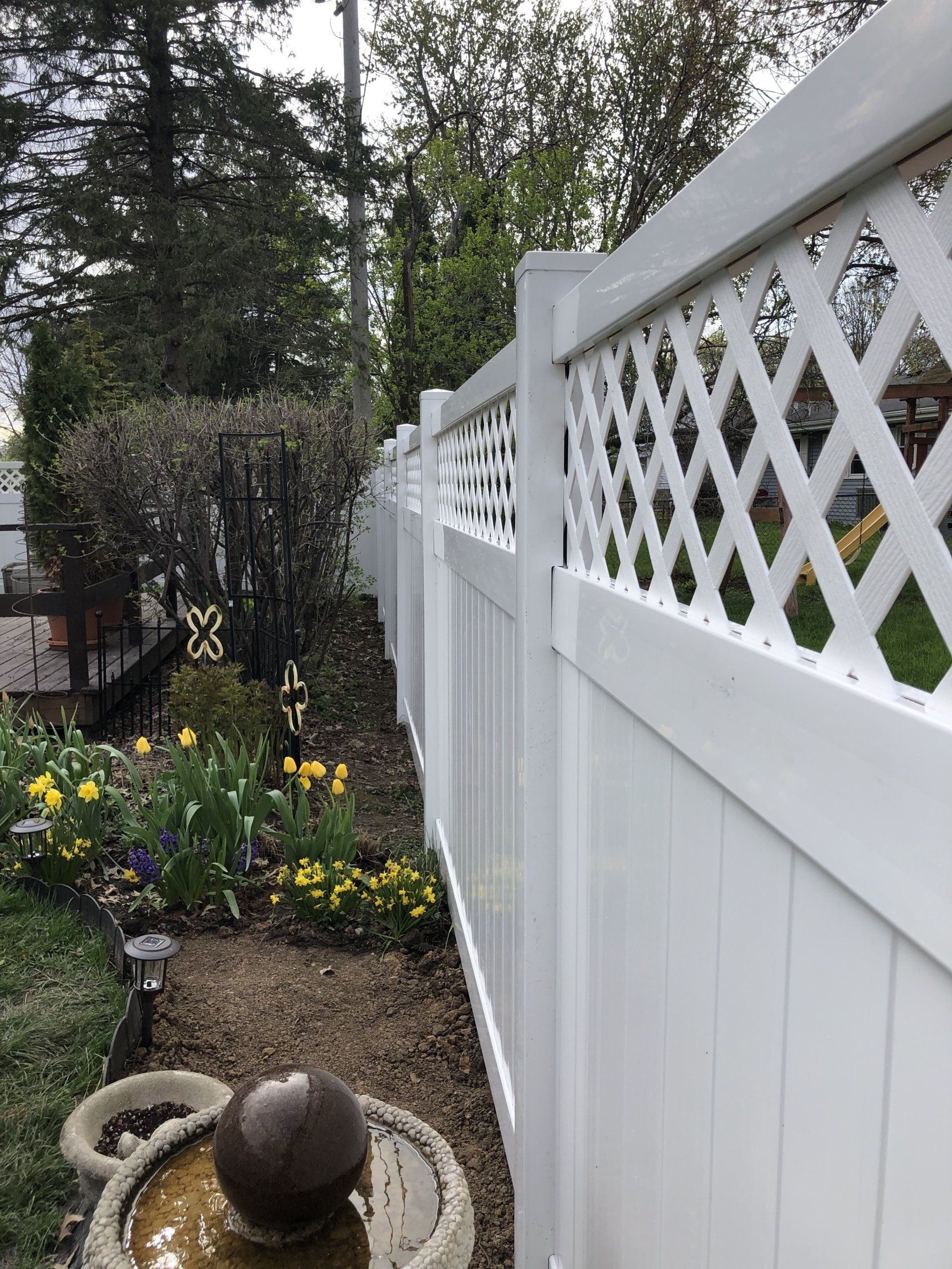 A white fence surrounds a garden with flowers and a fountain.