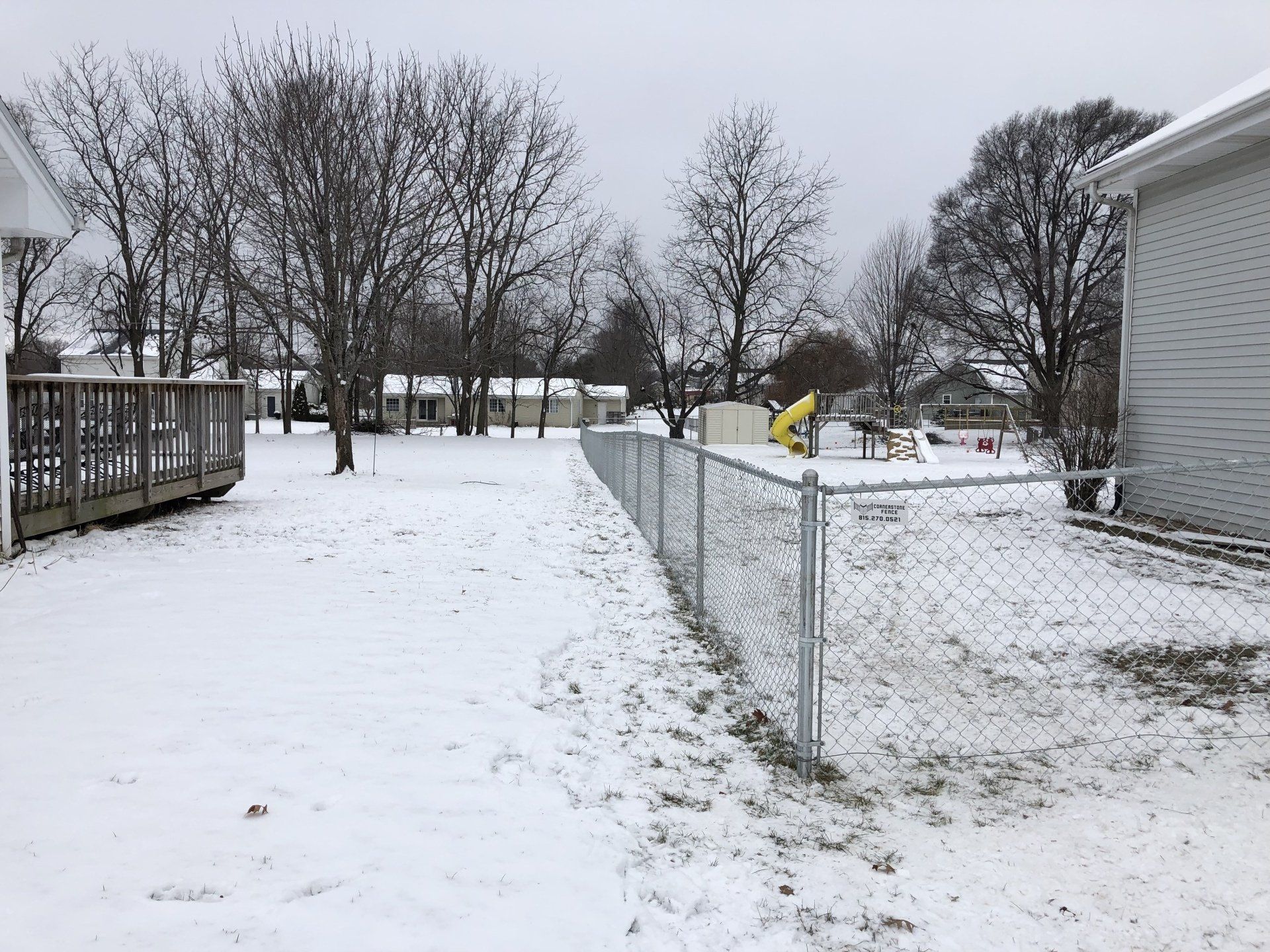 A snowy yard with a chain link fence and a house in the background.