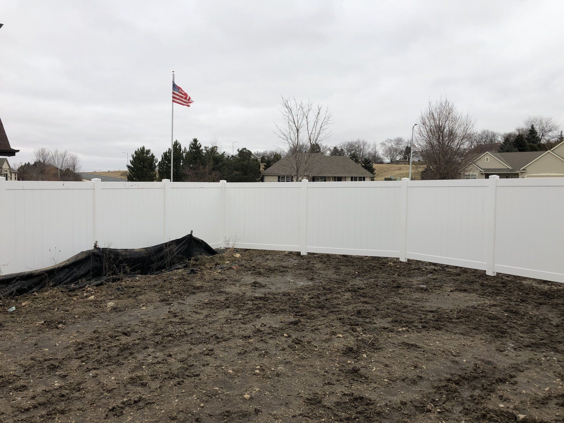 A white fence surrounds a dirt field with a flag in the background.