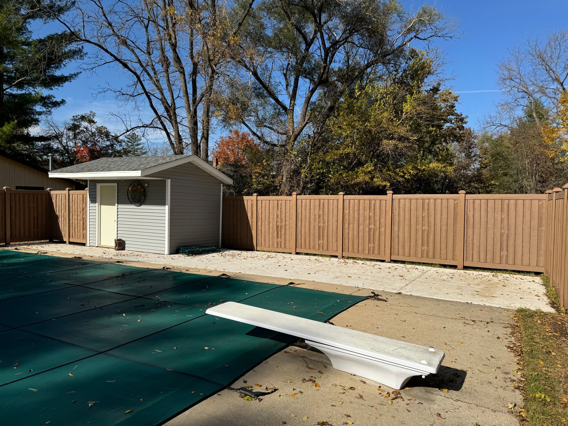 A wooden fence surrounds a swimming pool with a diving board in the foreground.