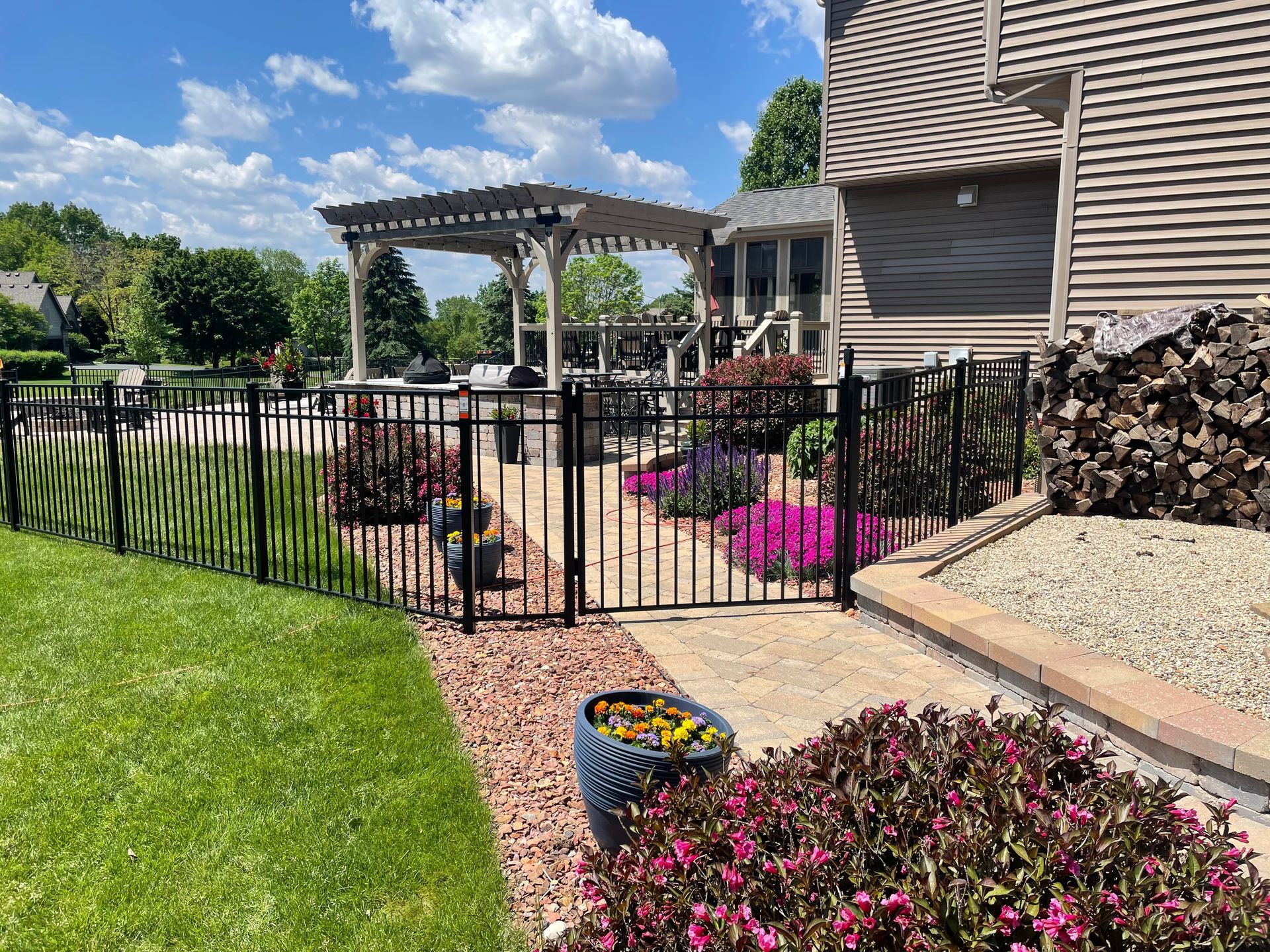 A black fence surrounds a patio with a pergola and flowers in front of a house.