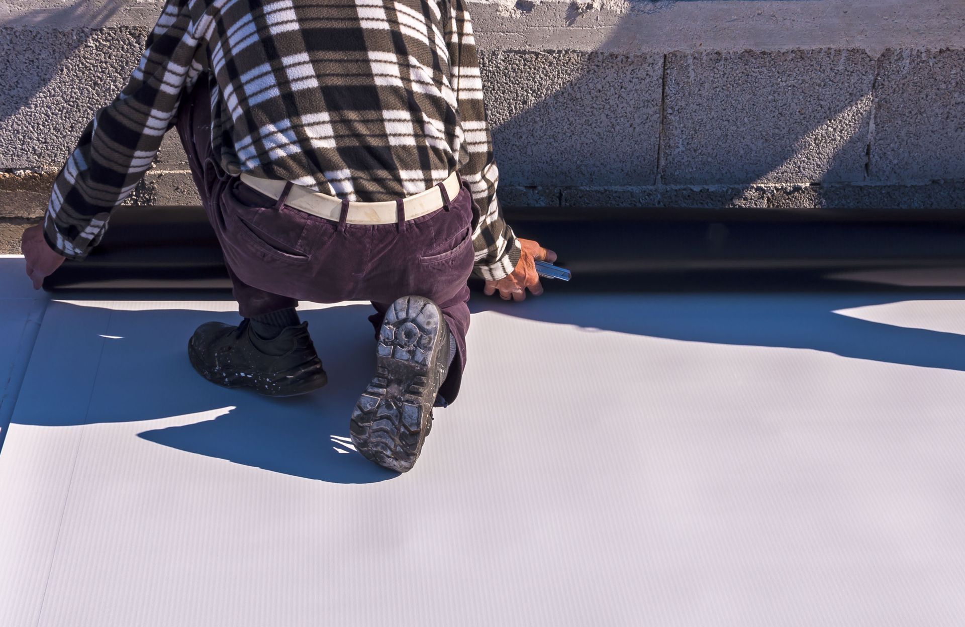Person kneeling, installing black sealant on white roofing material. Outdoor setting.