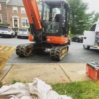 A small orange excavator is parked in a parking lot next to a sidewalk.