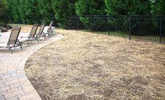 Three empty lounge chairs sit on a curved stone patio next to an area covered in straw mulch near a black metal fence.