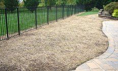 A patch of ground covered in light brown straw mulch next to a stone paver patio and a black metal fence.