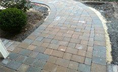 A curved walkway made of rectangular gray and brown pavers bordered by a darker stone edge, beside a green bush.