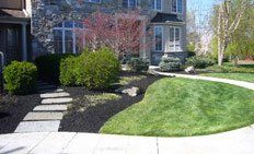 A stone pathway leads to a stone house with landscaped mulch beds, green shrubs, a small tree, and a curved lawn.