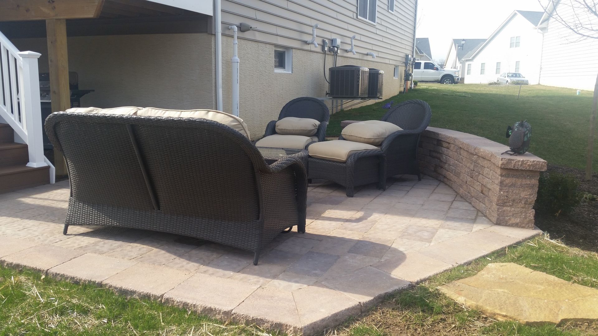 A patio with a brown wicker sofa and two matching chairs with beige cushions next to a low stone retaining wall.