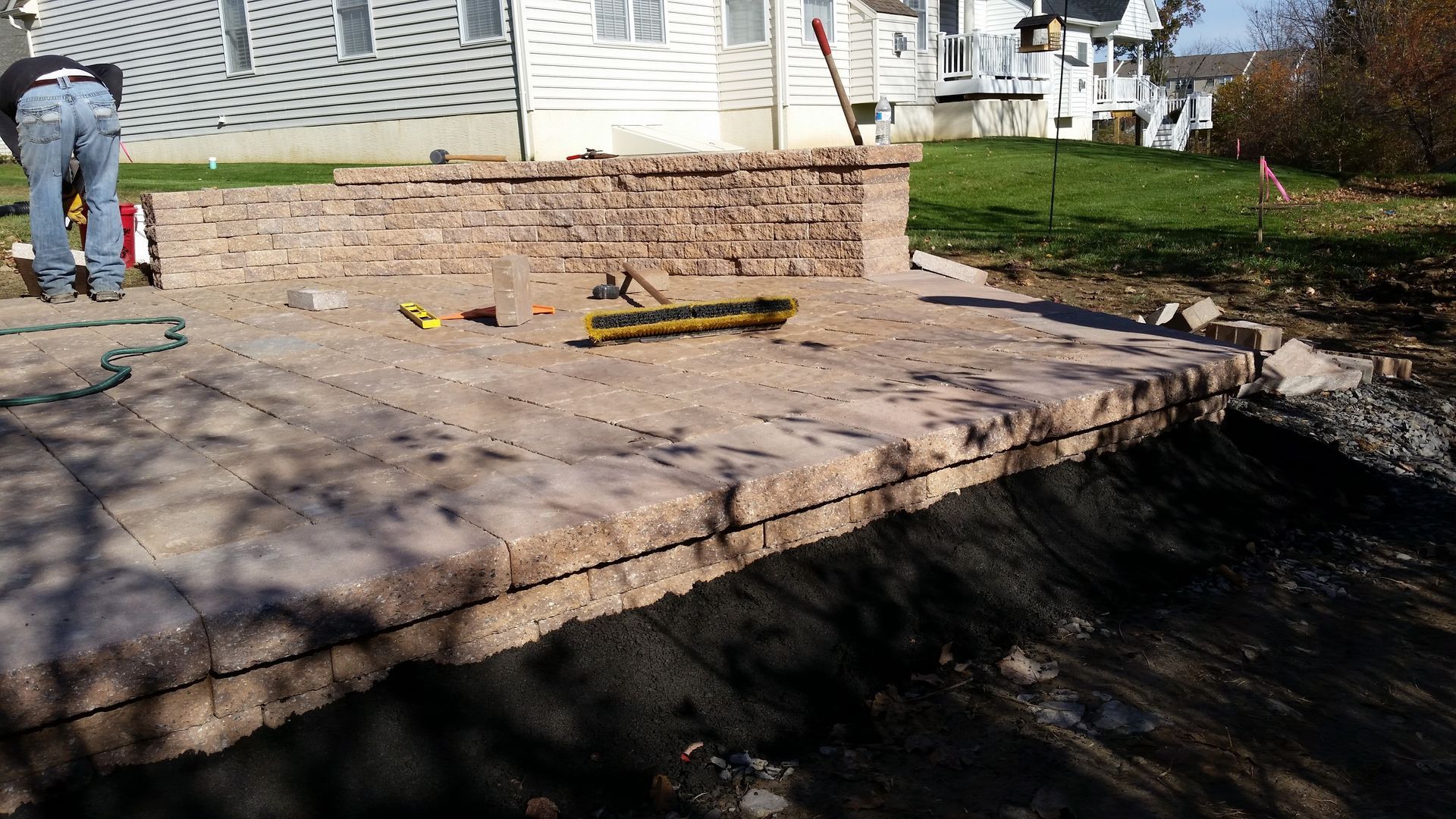 A person installing a tan stone patio and retaining wall in a grassy backyard.