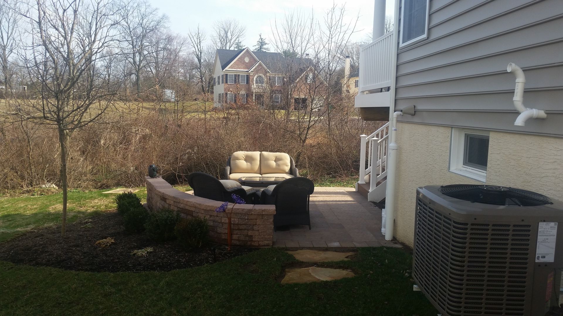 A backyard patio featuring a tan loveseat, two black chairs, and a curved brick sitting wall next to a house.