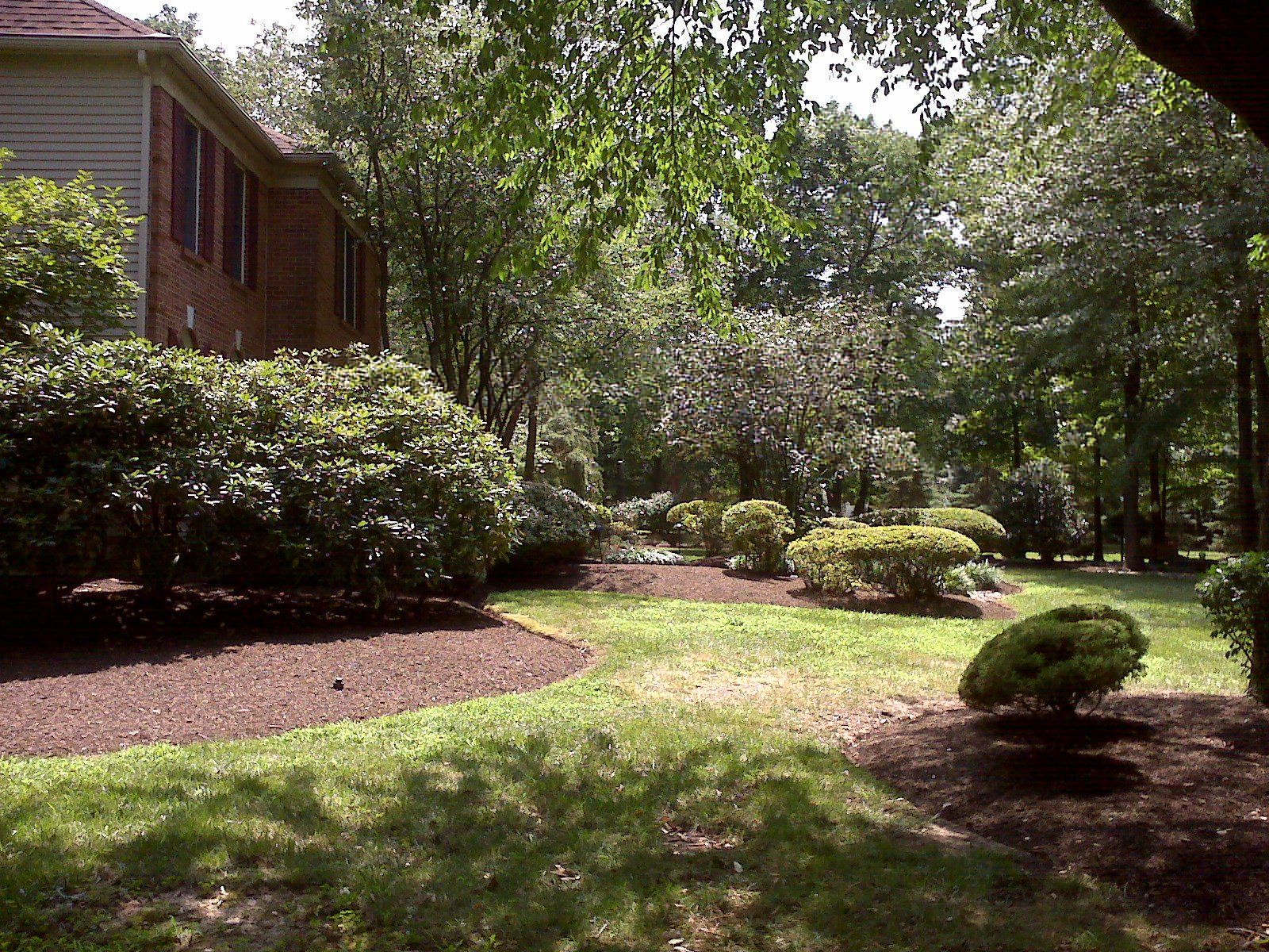A large brick and siding house sits beside a landscaped yard with mulched garden beds, shrubs, and trees in a sunny park.