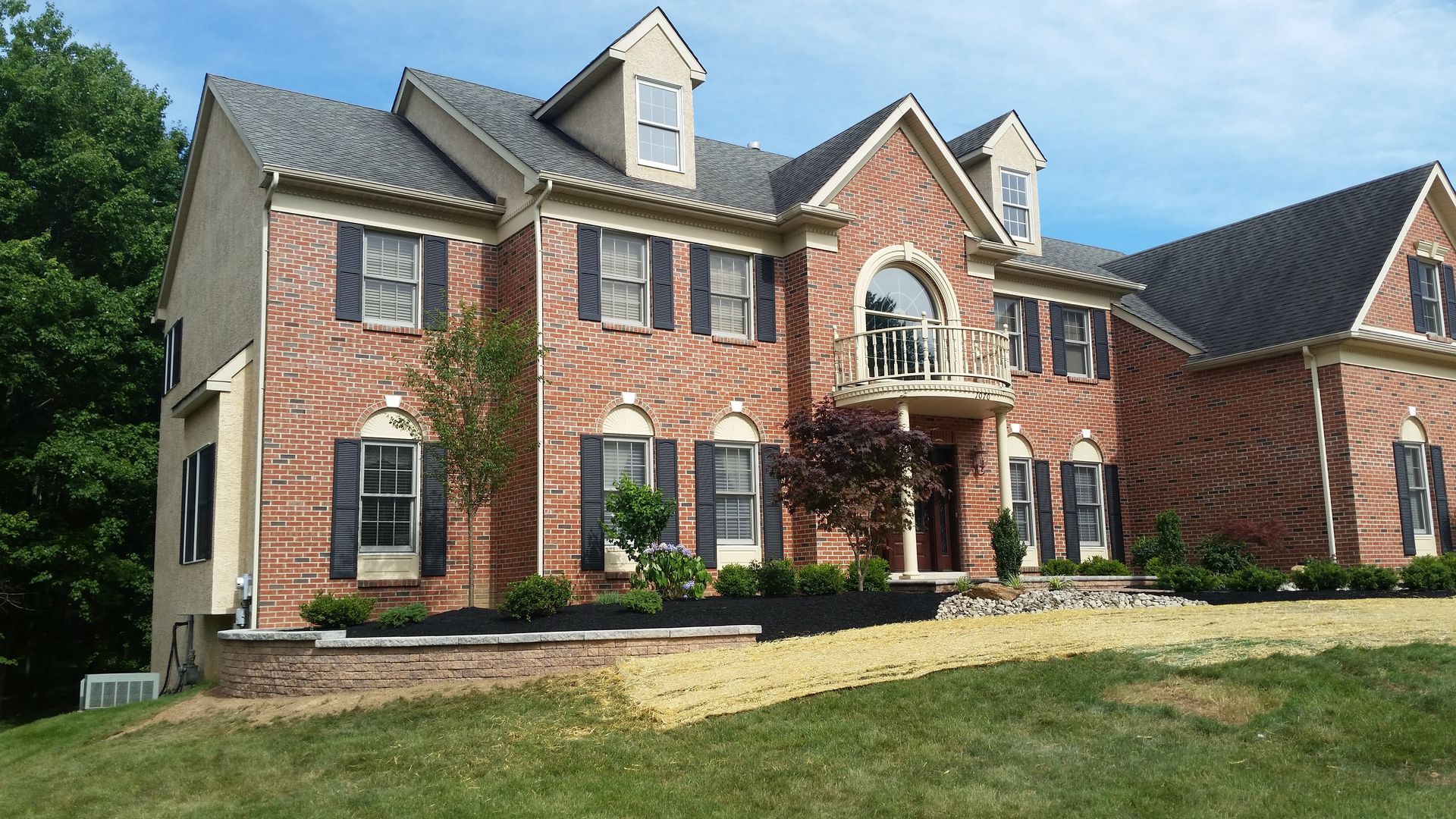 A two-story red brick house with dark shutters, a decorative arched balcony, and a manicured lawn under a blue sky.