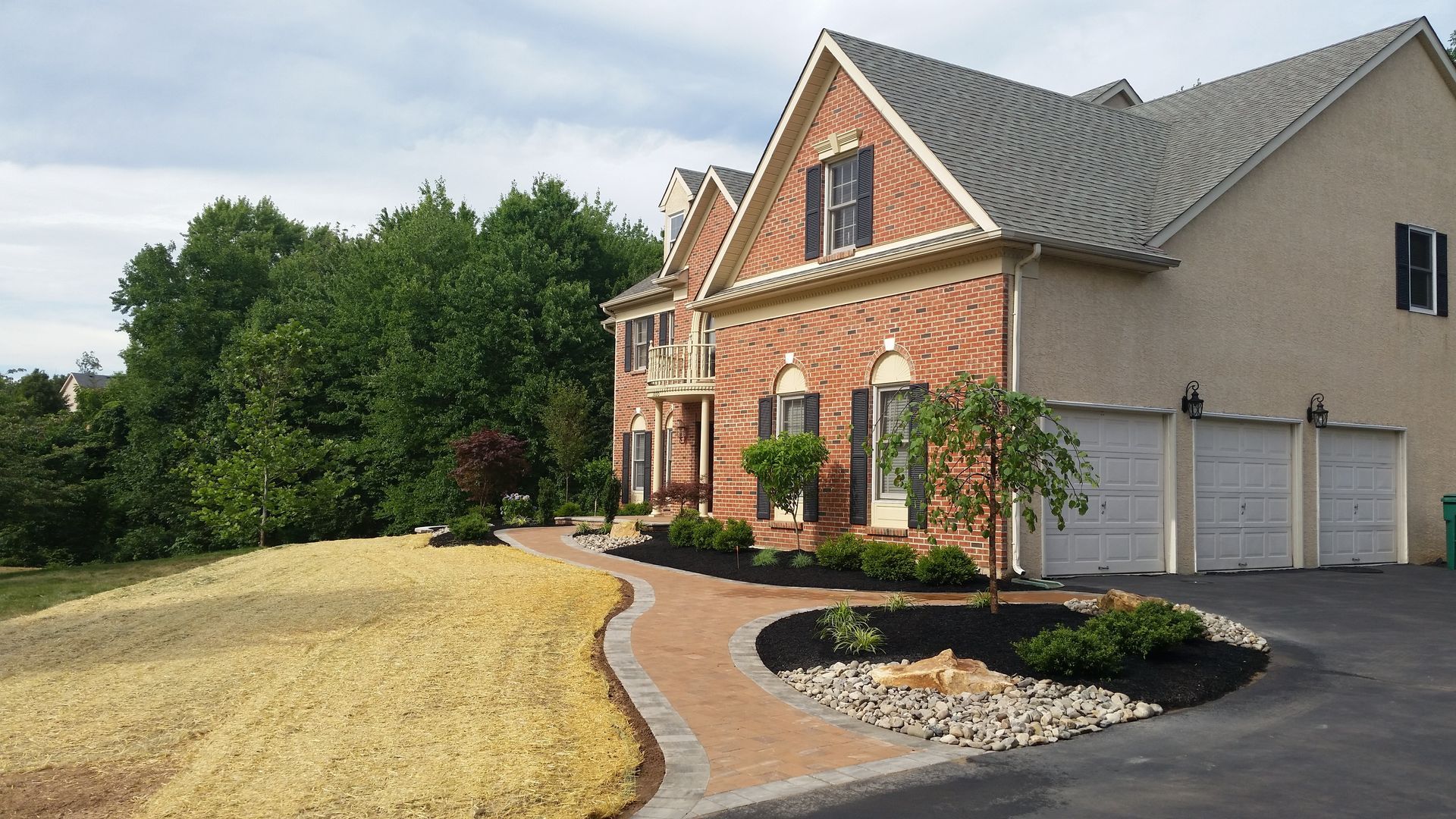 A two-story brick house with a paved walkway, landscaped garden beds, and a three-car garage next to a grassy lawn.