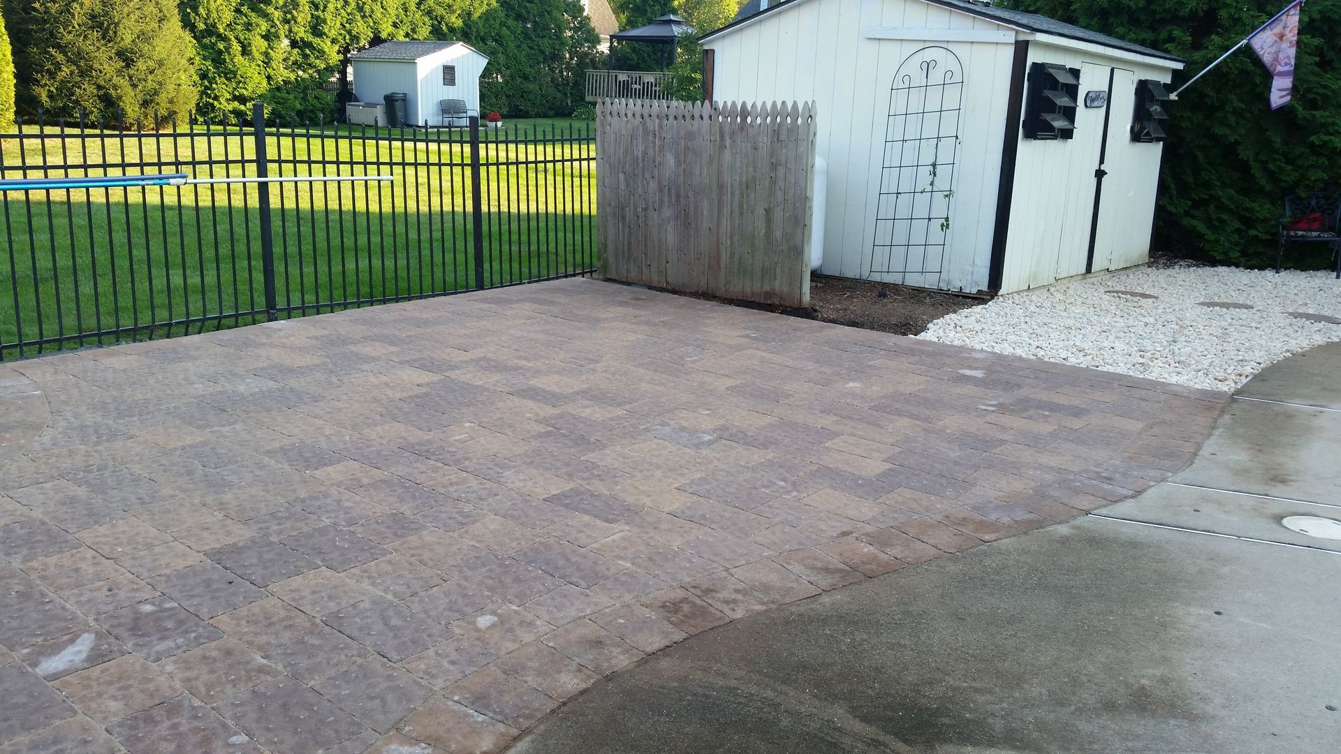 A brick patio sits in a backyard next to a small white shed, a wood privacy fence, and a green lawn.
