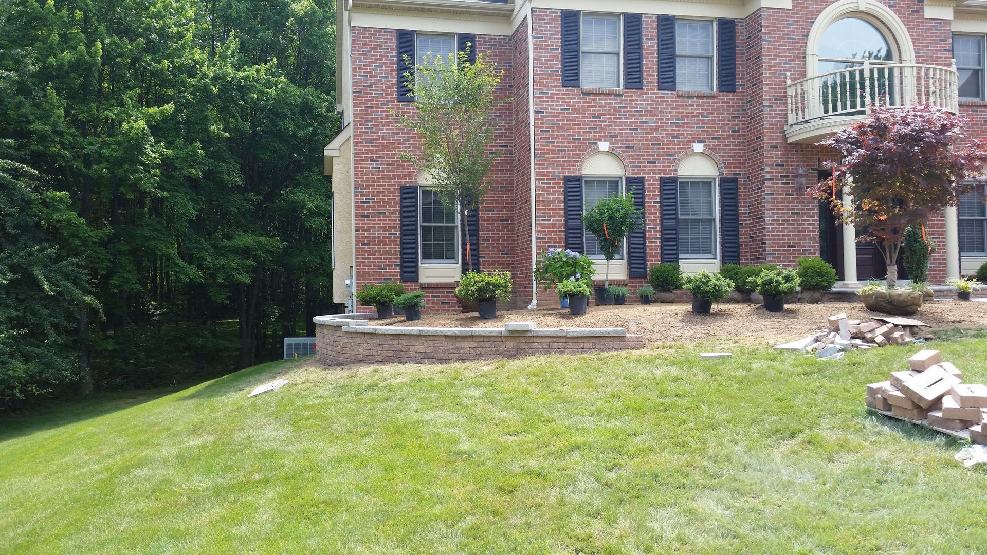 A brick two-story house with black shutters sits above a new stone retaining wall and landscaped garden beds.