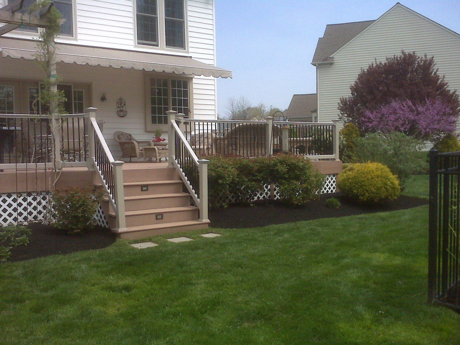 A white house with a tan deck and stairs leads down to a landscaped backyard with green lawn, mulch, and shrubs.