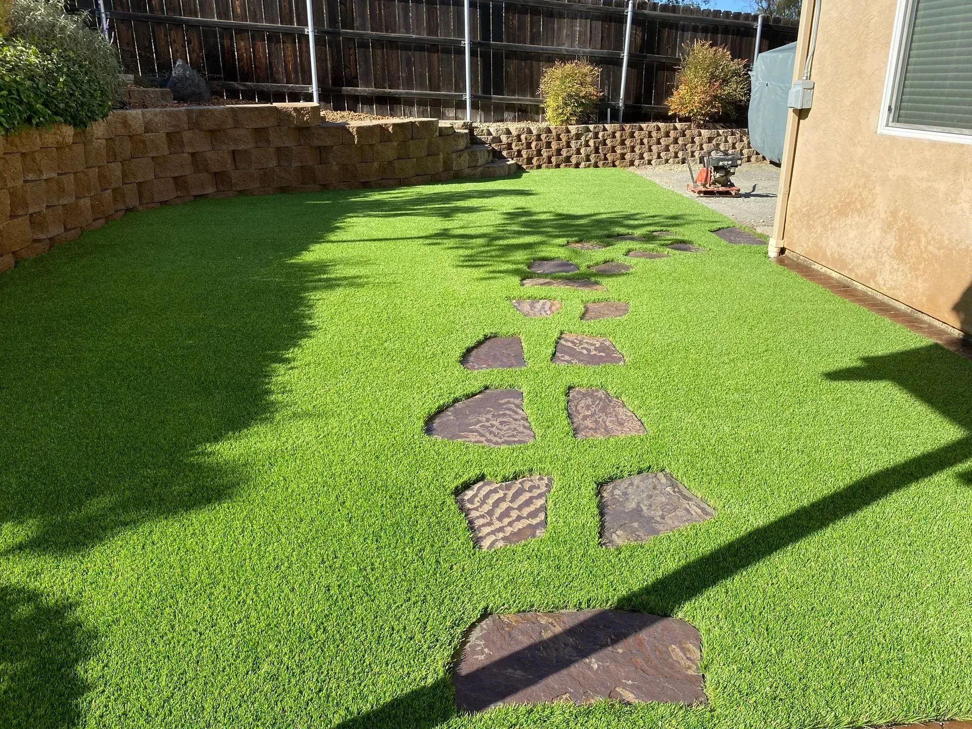 Green artificial lawn with stone stepping stones and retaining wall in a backyard.