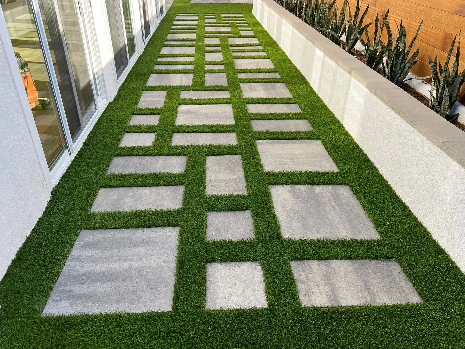 Gray stepping stones on green turf, along a building with a white wall and plants.