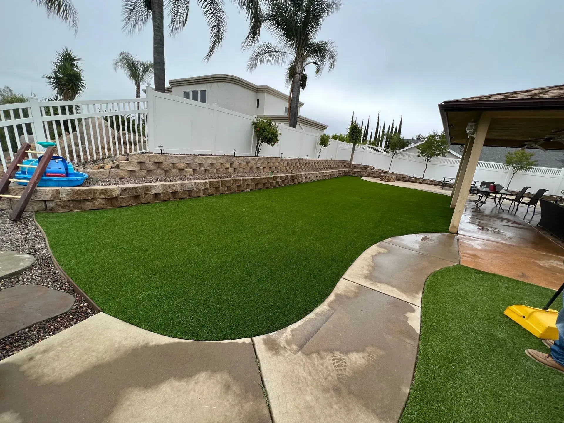 Green artificial turf backyard with pathway, retaining wall, and covered patio.