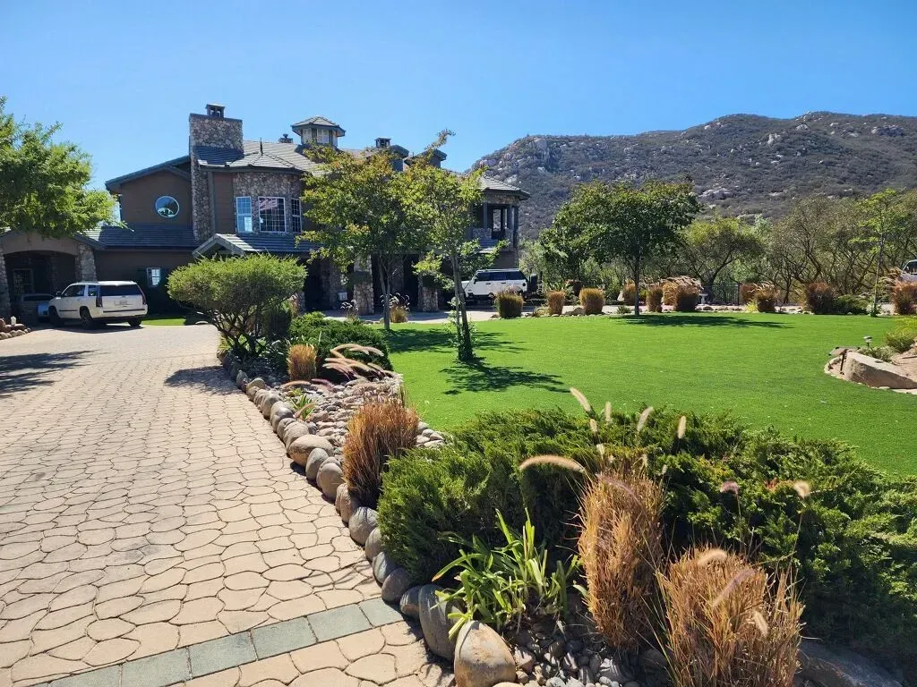 Large stone house with circular driveway, green lawn, trees, and mountain backdrop.