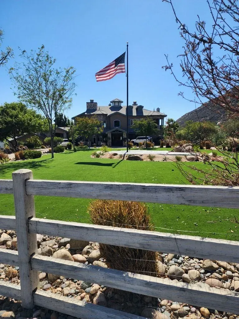 Large house with American flag, green lawn, and white fence under a blue sky.