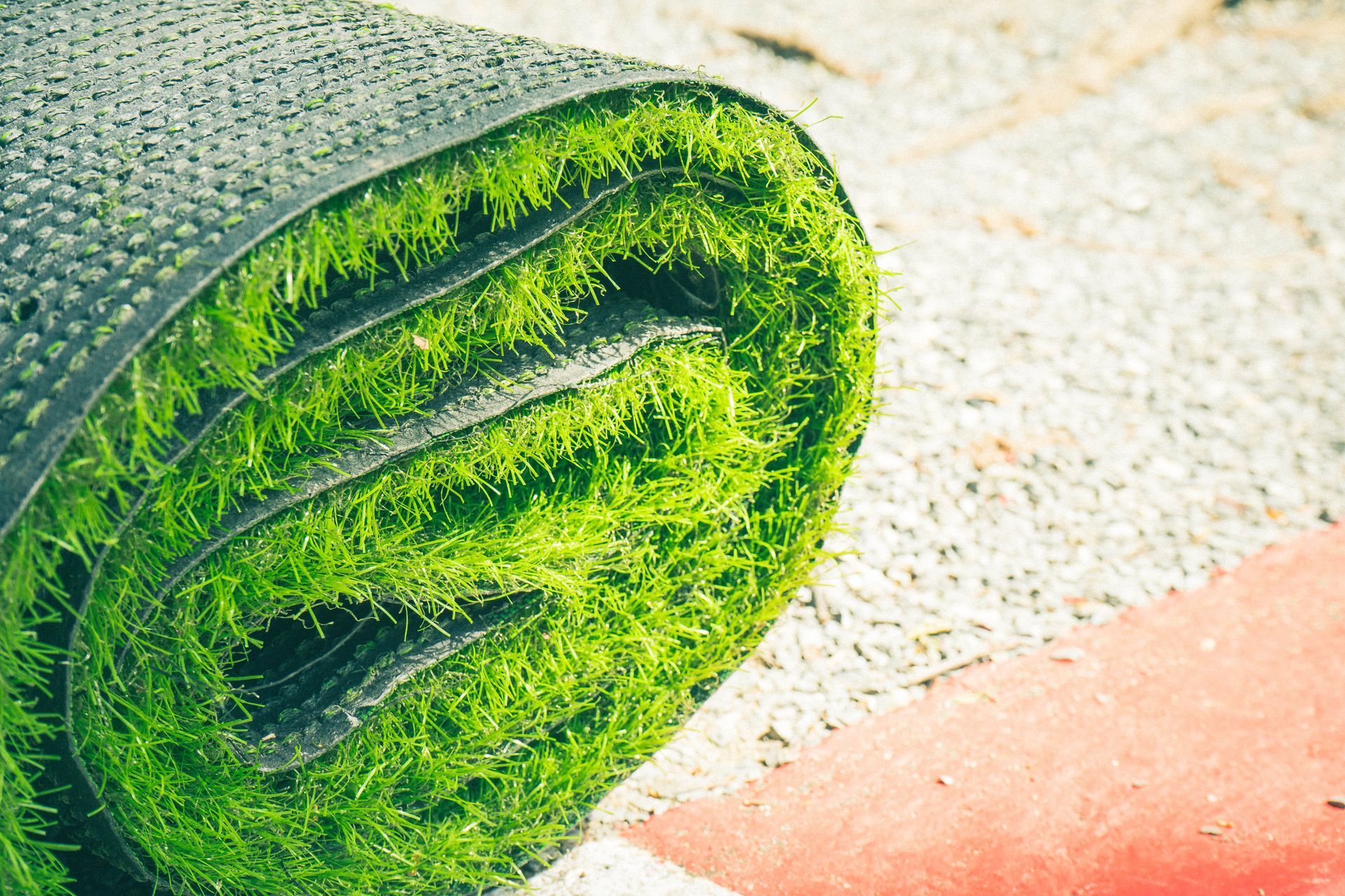 Rolled-up artificial turf, showing green blades and black backing, on a gravel surface.