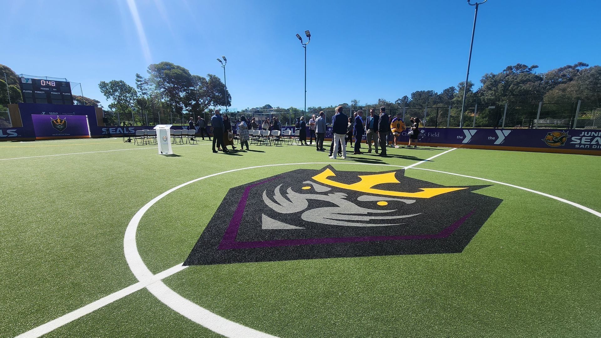 Soccer field with a team logo in the center, people standing around, sunny day.