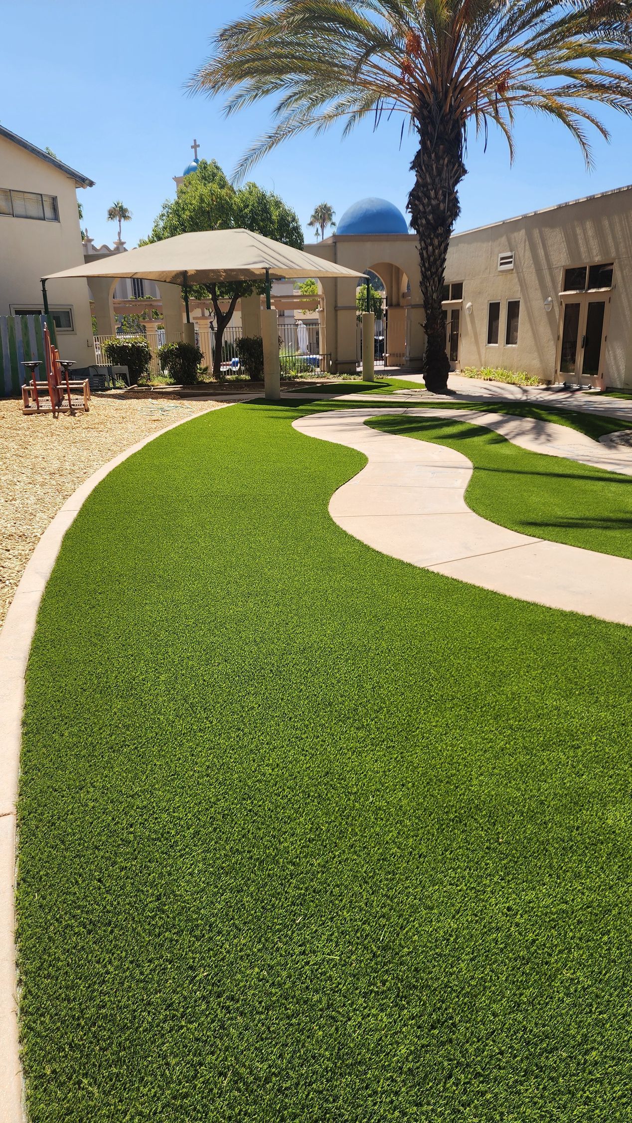 Lush green lawn with a winding sidewalk, surrounded by buildings and a gazebo under a sunny sky.