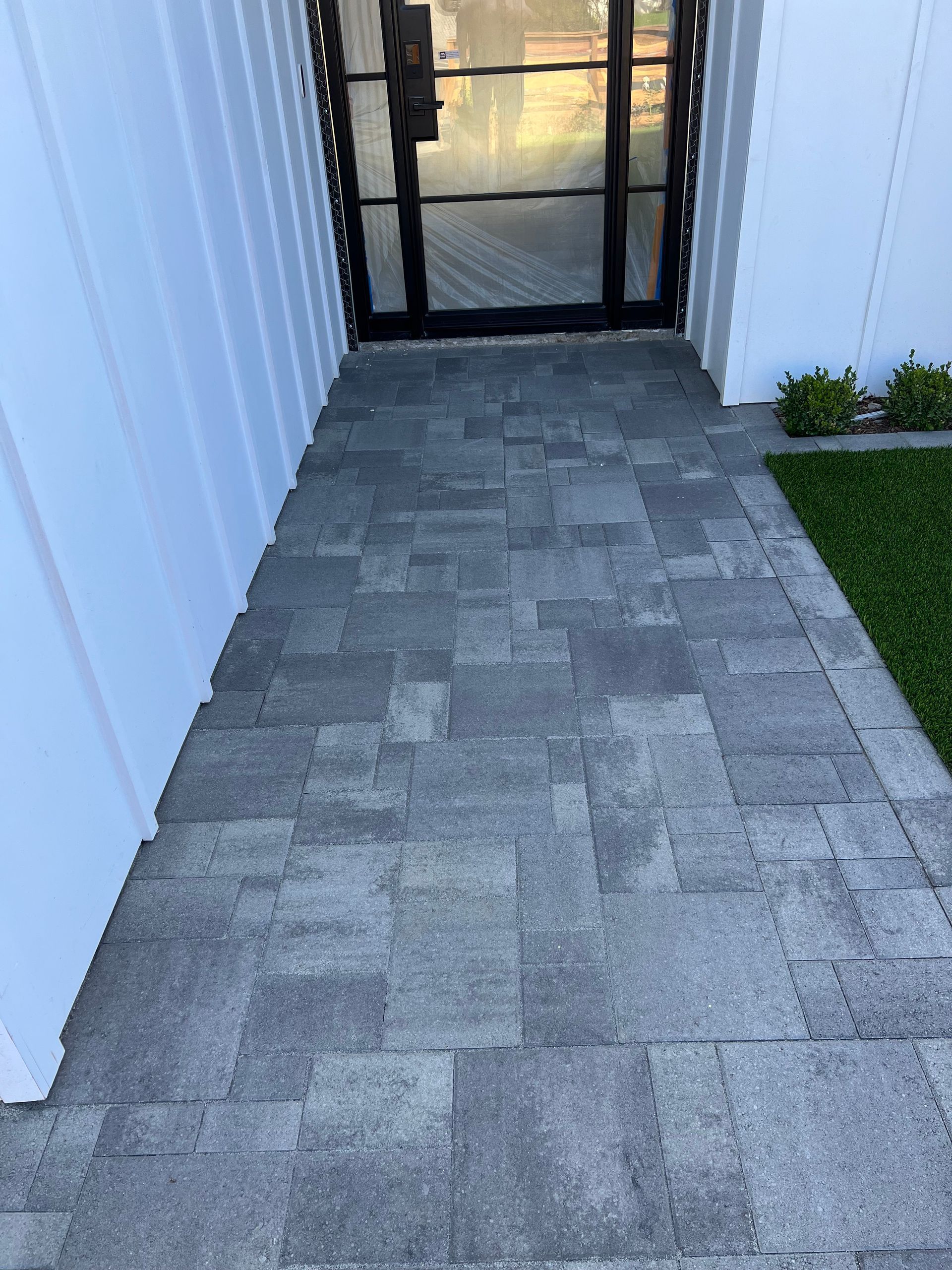 Gray stone walkway leading to a black-framed glass door, with white siding on the left and artificial turf on the right.