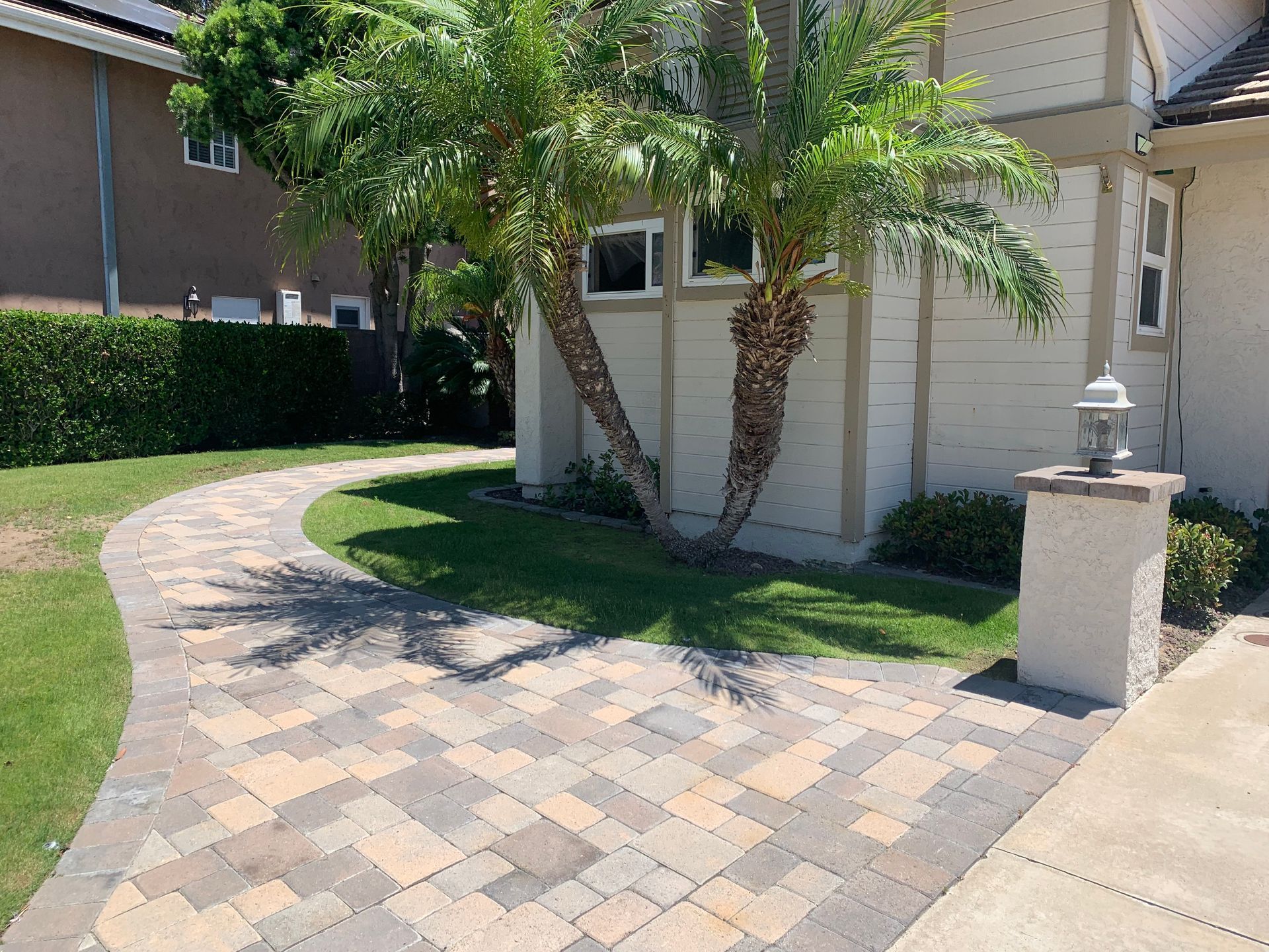 Brick pathway curves toward a house with palm trees, green grass, and a decorative pillar.