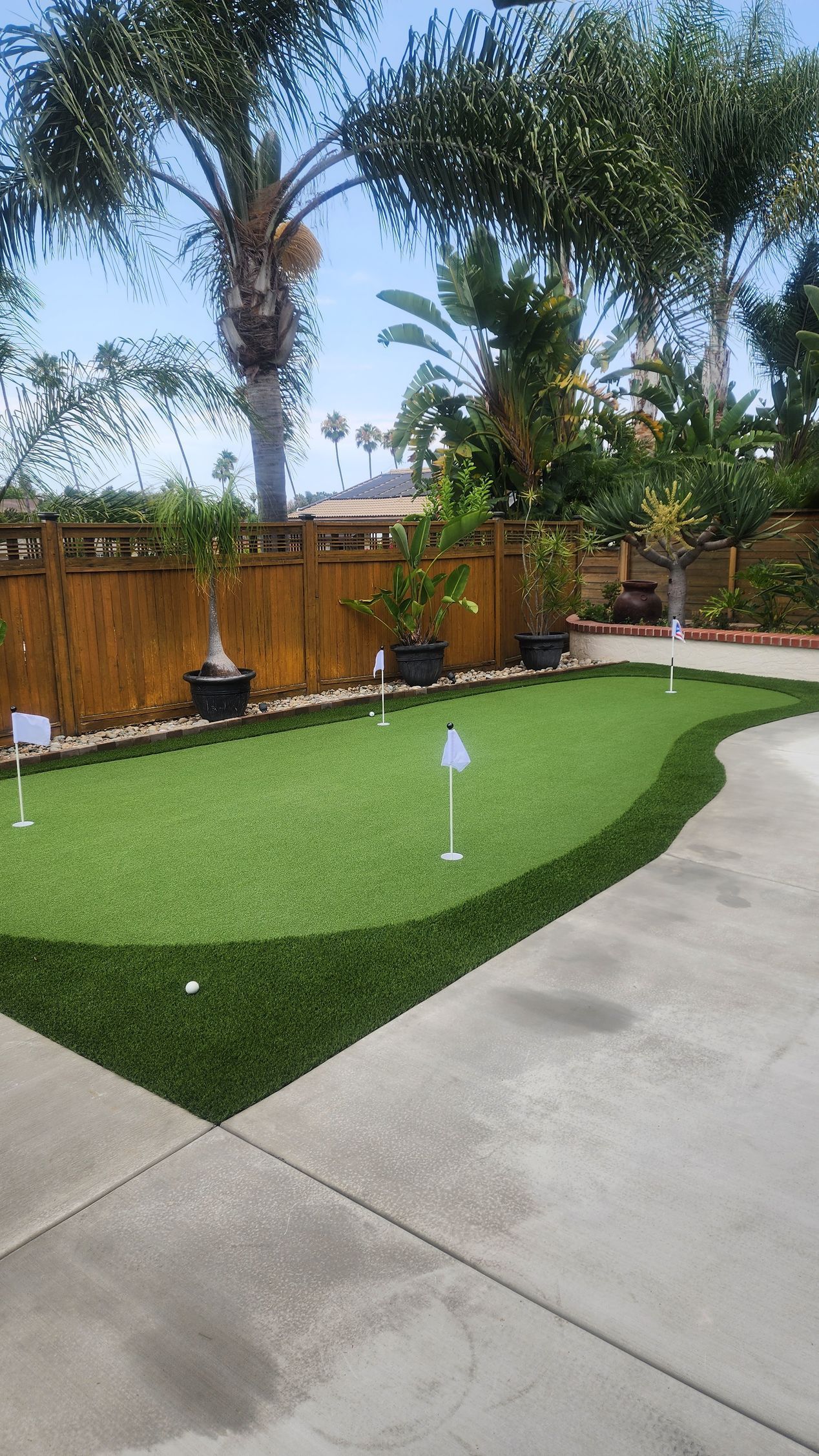 Backyard putting green with flags, surrounded by concrete and wooden fence, under blue sky.